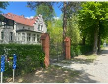 Haus mit weißer Fassade und rotem Ziegeldach, flankiert von einem Metalltor mit Backsteinpfeilern. Der umliegende Garten ist mit Hecken und hohen Bäumen umgeben, ein gepflasterter Gehweg verläuft parallel zur Straße.