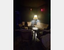 Sitzecke in einem dunklen Raum mit warmem Licht; ein Sofa und mehrere Sessel umgeben zwei kleine runde Tische, beleuchtet von einer Stehlampe mit einem plissierten Lampenschirm. An den dunklen Wänden sind zwei kleine Wandlampen mit rotem Licht angebracht.