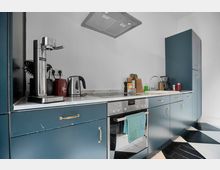 Moderne Küche mit blauen Fronten und goldfarbenen Griffen. Zu sehen sind ein Herd mit Backofen, eine Dunstabzugshaube, eine Spüle und eine Arbeitsplatte mit Haushaltsgeräten wie Wasserkocher und Sprudler; der Boden ist mit einem schwarz-weißen geometrischen Muster gestaltet.