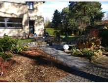 Garten mit gepflastertem Weg, Steinmauern, mehreren Pflanzenbeeten und einem großen Baumstumpf als Dekoration. Links ein Wohnhaus mit Putzfassade, im Hintergrund Bäume, Rasen und eine Gartenlaube.