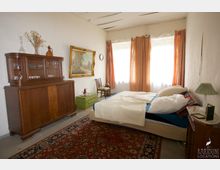 Ein Schlafzimmer mit schlichter Einrichtung: Ein Doppelbett mit weißer Bettwäsche steht rechts, daneben ein kleiner Nachttisch mit Lampe. Links ein Holzbuffet, darauf Dekoration und Geschirr. Ein gemälde in einem goldenen Rahmen hängt an der Wand, die Fenster sind mit orangefarbenen Vorhängen versehen, und ein großer Teppich bedeckt den Boden.