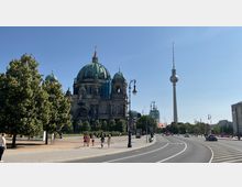 Straßenansicht in Berlin mit dem Berliner Dom auf der linken Seite, dessen grüne Kuppel von Bäumen umgeben ist, und dem Fernsehturm im Hintergrund rechts. Die Szene zeigt eine breite Straße mit Gehwegen und Straßenlaternen bei sonnigem Wetter.