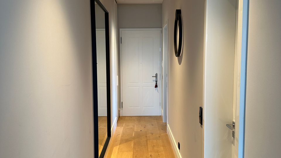 Schmaler, moderner Flur mit hellgrauen Wänden, hellem Holzboden und weißer Decke mit schwarzer Deckenleuchte. Links hängt ein großer Wandspiegel, rechts ist eine offene Türöffnung; am Ende des Gangs befindet sich eine weiße Zimmertür.