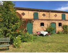 Altes Backsteingebäude mit grünen Holztüren und Fensterläden, inmitten eines Gartens mit Grasfläche, Sträuchern und einem kleinen Tisch mit Tischdecke sowie zwei Stühlen. Links steht eine einfache Holzgartenbank unter einem Baum.