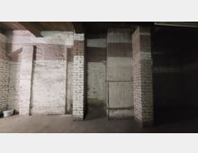 Kellerraum mit weißen Ziegelwänden, betonierten Boden und sichtbaren Holzbalken an der Decke. Die Wand ist durch vertikale Pfeiler unterteilt, und der Raum wirkt dunkel und spärlich beleuchtet.