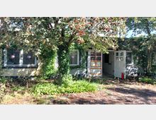 Eingewachsenes Gebäude mit cremefarbener Fassade und großen Fenstern, teilweise mit Efeu bedeckt. Im Vordergrund steht ein Baum mit roten Beeren, und der Eingang mit halb geöffneter Tür ist sichtbar; daneben ein Feuerlöscher.