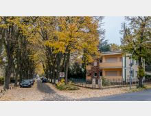 Straßenszene in einer Wohngegend mit herbstlichen Bäumen, die die Straße säumen. Links geparkte Autos, rechts moderne Wohnhäuser mit gelblichen und braunen Fassaden; ein Straßenschild zeigt "Auguststraße", und der Boden ist mit fallendem Laub bedeckt.