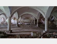 Unterirdischer Raum mit einer Reihe von gemauerten Rundbögen, die von steinernen Säulen gestützt werden. Der Raum wirkt verlassen und enthält verstreute Trümmer sowie verwitterte Wandstrukturen aus Ziegeln.