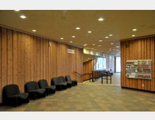 Holzvertäfelter Innenraum eines öffentlichen Gebäudes mit modernen schwarzen Sitzmöglichkeiten entlang der Wand, einer Garderobenbeschilderung und einem Regal mit Informationsbroschüren auf der rechten Seite. Im Hintergrund befindet sich ein Flur mit Glaswänden, der zu einem Ausgang führt.