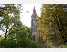 Kirchturm eines historischen Backsteingebäudes, teilweise verdeckt von hohen, dicht belaubten Bäumen, in einem Außenbereich bei Tageslicht.