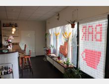 Eine kleine Bar mit Tresen, Holzhockern und dahinterliegendem Regal mit Gläsern und Flaschen. Rechts befindet sich eine Fensterfront mit transparenten Vorhängen und Pflanzendekoration, durch die ein Schild mit dem Schriftzug "BAR" sichtbar ist. Im Hintergrund eine Tür mit der Aufschrift "WC".