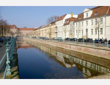 Eine Kanallandschaft in einer städtischen Umgebung mit historischen Gebäuden im Hintergrund. Der Kanal ist von einem Gehweg und einer grünen Geländerreihe gesäumt, entlang derer Autos geparkt sind.