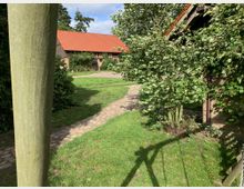 Ländlicher Hof mit gepflasterten Wegen, grünem Rasen und Bäumen. Im Hintergrund ein Gebäude mit rotem Ziegeldach, im Vordergrund ein Ausschnitt eines Holzpfostens und einer Backsteinmauer.