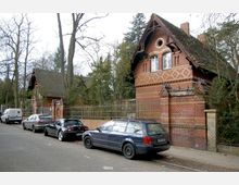 Straßenszene mit parkenden Autos vor einem historischen Backsteingebäude mit verziertem Mauerwerk, Spitzdach und schmiedeeisernem Zaun, umgeben von hohen Bäumen. Die Umgebung wirkt wie ein ruhiges Wohn- oder Parkgebiet.