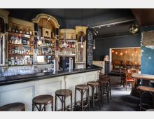 Innenansicht einer gemütlichen Bar mit einer langen Theke aus Holz, Barhockern und Regalen voller Flaschen und Gläser. Im Hintergrund befindet sich ein Bereich mit Sitzplätzen, Holzstühlen und warmem, gedämpftem Licht an den Wänden.