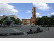 Weitläufiger Platz mit einem Brunnen im Vordergrund, der Figuren und Wasserspiele zeigt, sowie dem roten Backsteingebäude des Berliner Rathauses im Hintergrund; umgeben von Bäumen und vereinzelten Menschen auf Sitzbänken.