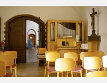 Innenraum einer Kirche mit einem kleinen Versammlungsraum. Im Vordergrund befinden sich Holzstühle, daneben steht eine helle Orgel, und an der Wand ein Kruzifix. Im Hintergrund führt eine geöffnete Tür zu einem gewölbten Durchgang mit Backsteinakzenten.