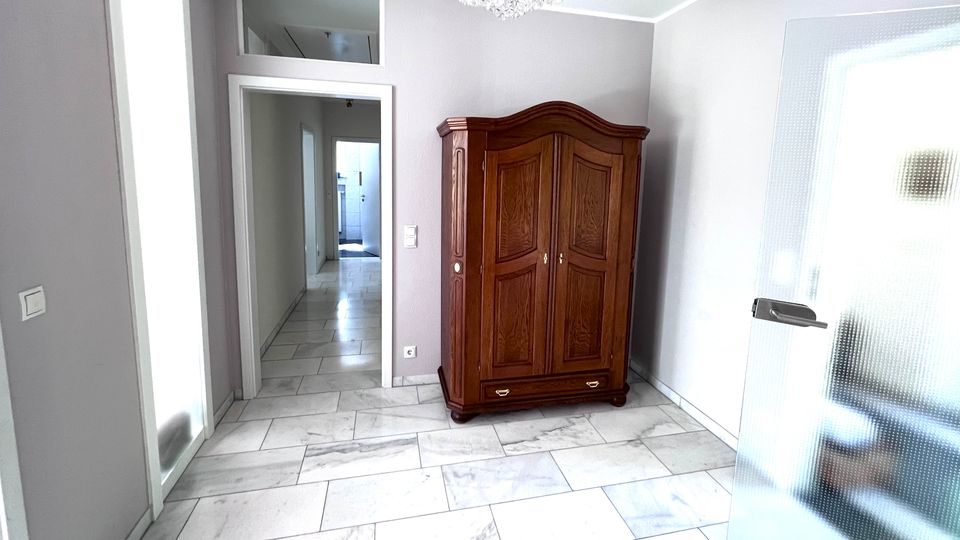 Heller Eingangs- oder Flurbereich mit hellgrauen Wänden und großformatigem, marmoriertem Fliesenboden. In der Ecke steht ein dunkler Holzschrank; darüber hängt ein Kristalllüster, daneben führt eine Tür mit Milchglas in den Raum und ein offener Durchgang in einen langen Flur.
