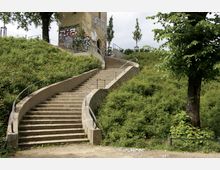Eine geschwungene Steintreppe mit Metallgeländern führt einen grasbewachsenen Hang hinauf zu einem Gebäude aus gelbem Stein, das mit Graffiti bedeckt ist. Umgeben von Bäumen und Büschen vermittelt die Szene einen parkähnlichen Eindruck.