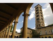 Eine historische Kirche mit einem rechteckigen Glockenturm aus Backstein, umgeben von einem Arkadenhof mit gelblichen Säulen und dekorativen Kapitellen. Im Hintergrund ist ein teilweise bewölkter Himmel zu sehen.