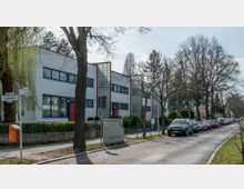 Moderne Reihenhäuser im Bauhausstil mit weißen Fassaden, blauen Fensterrahmen und roten Türen, ergänzt durch vertikale Glasbausteine. Im Vordergrund eine Straße mit parkenden Autos, Gehwegen und großen Bäumen.