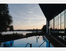 Außenpool mit mehreren Personen, gelegen an einem See mit umliegender Vegetation. Rechts im Bild spiegelt sich der Sonnenuntergang in einer modernen Glasfassade, während im Hintergrund Boote und eine ruhige Landschaft zu sehen sind.