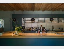 Moderne Bar mit einer Theke aus Holz und hängenden schwarzen Lampen, flankiert von mit Flaschen, Gläsern und Geräten bestückten Regalflächen vor einer hellen Rückwand. Die Decke besteht aus sichtbaren Holzbalken, und links ist ein Fenster mit abgerundeten Bögen sichtbar.