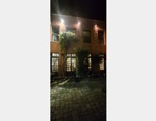 Nächtlicher Innenhof mit gepflastertem Boden vor einem zweigeschossigen Gebäude mit rau verputzter Fassade, mehreren Fenstern und großflächigen Glas-Türen im Erdgeschoss. Außenleuchten beleuchten die Wand, und Kletterpflanzen sowie große Pflanzkübel stehen entlang der Fassade.