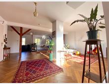 Wohnzimmer in einem offenen, hellen Raum mit Holzboden im Fischgrätmuster, mehreren Teppichen und Holzbalken. Die Einrichtung umfasst Pflanzen, einen Tisch, Stühle und eine offene Küche im Hintergrund vor großen Fenstern.