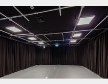 Ein rechteckiger schwarzer Raum mit vollständig von schwarzen Vorhängen bedeckten Wänden und einer dunkelgrauen Bodenfläche. An der Decke befinden sich Metallträger mit rechteckigen LED-Leuchten und technischem Equipment.