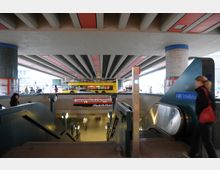 U-Bahnhofeingang „Schloßstraße“ in Berlin, mit Rolltreppe und Treppenabgang. Im Hintergrund eine mehrspurige Straße mit einem gelben Doppeldeckerbus, der unter einer breiten Betonbrücke fährt.