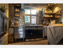 Rustikale Küche mit dunklen Fliesenwänden, großem Gaskochherd mit goldenen Details, darunter eine gemauerte Arbeitsfläche und ein Fenster mit Blick ins Freie. Regale mit Gewürzen und Küchenutensilien sowie ein Korb mit Holzlöffeln ergänzen die Einrichtung.