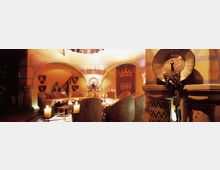 Ein mittelalterlich gestalteter Raum mit einem runden Tisch, umgeben von hohen Stühlen. Die Wände sind mit Wappen, Rüstungen und einem großen Wandteppich mit einer Krone verziert, während Kerzen den Raum beleuchten.