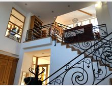 Innenansicht eines Wohnhauses mit einer zweistöckigen Treppe aus Holzstufen und dekorativen schmiedeeisernen Geländern. Im Hintergrund sind große Fenster, eine Galerie mit Bücherregal und eine hängende Deckenleuchte zu sehen.