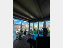 Wintergarten mit Holzfußboden und Glaswänden, der Blick auf eine Dachterrasse mit Pflanzen und Sitzmöbeln freigibt; im Hintergrund sind Dächer und Gebäude einer Stadt zu sehen.