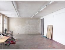 Ein Künstleratelier mit einer großen weißen und einer beige-fleckigen Wand, einem Boden mit Farbspritzern und hohen Fenstern auf der linken Seite. Im Raum stehen ein Arbeitstisch mit Kunstmaterialien, ein beweglicher Regalwagen und eine gestützte Leinwand an der Wand.