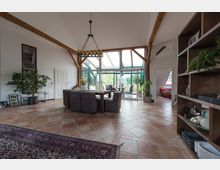 Wohnzimmer mit hohen Decken, sichtbaren Holzbalken und großen Glasfenstern, die auf eine Terrasse führen. Der Raum ist mit Fliesenboden, einem Esstisch und einer Bücherwand ausgestattet, dekoriert mit Pflanzen und Wohnaccessoires.