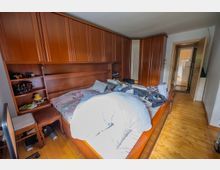 Schlafzimmer mit großem Holzbett und wandfüllendem Holzschrank, der ein integriertes Regal und eine Ablage am Kopfteil bildet. Holzdielenboden und offene Tür zu einem Flur mit Blick in einen weiteren Raum.