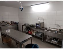 Eine gewerbliche Küchenarbeitsfläche mit weißen gefliesten Wänden, Edelstahl-Arbeitsplatten, einer Spüle, verschiedenen Geräten wie einer Vakuumverpackungsmaschine und einer Schneidemaschine sowie Regalen mit Zubehör und Utensilien. Die Beleuchtung erfolgt durch eine Deckenleuchte.
