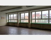 Leerer, älterer Innenraum mit großen Fensterfronten entlang einer Wand, grünen Fensterbänken und Heizkörpern darunter. An der Decke hängen lange Leuchtstofflampen; durch die Fenster sind Wohngebäude und Bäume zu sehen.