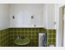 Ein kleines Badezimmer mit weißen Holzpaneelen an den Wänden und grün-gelber Retro-Keramikfliesen mit kreisförmigem Muster im unteren Bereich. Es gibt ein grünes Waschbecken, einen alten, weißen Heizkörper, eine Ablage mit einer Toilettenpapierrolle, zwei kleine Wandlampen und ein gestreiftes Handtuch, das an der Seite hängt.