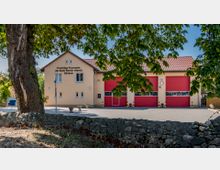 Außenansicht eines Feuerwehrhauses der Freiwilligen Feuerwehr Glindow, Stadt Werder (Havel), mit beiger Fassade, drei großen roten Toren und einem roten Ziegeldach; im Vordergrund ein gepflasterter Hof und ein Baum mit Steinmauer.