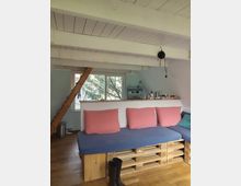 Innenraum eines kleinen Dachgeschoss-Studios mit weiß gestrichenen Holzbalken und niedrigem Plafond, großem Fenster und einer einfachen Küchenzeile im Hintergrund. Im Vordergrund steht ein Paletten-Sofa mit blauem Polster und rosa Kissen auf Holzboden, daneben eine schräge Holztreppe und eine hängende Lampe.