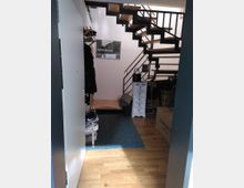 Eingangsbereich eines Hauses mit Holzfußboden, einer offenen Holz- und Metalltreppe, einer Garderobe an der Wand, einem blauen Teppich und einem Regal mit Schubladen. Im Hintergrund ist eine Wanddekoration mit einem Foto erkennbar.