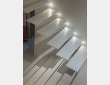 Interior view of a modern staircase with white floating rectangular steps attached to a wall, lit by small round recessed lights along the side. The steps cast strong linear shadows onto the lower landing and wall.