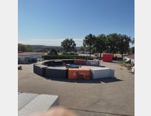 Ein großer, offener Platz mit Containern, die in einem unregelmäßigen Kreis angeordnet sind. Im Hintergrund sind Bäume, ein kleineres Gebäude und ein weiter Himmel zu erkennen.