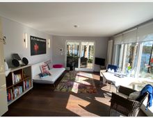 Modernes Wohnzimmer mit heller Einrichtung, großer Fensterfront und Blick auf Bäume und Wasser. Der Raum enthält ein Sofa, einen kleinen Esstisch, Bücherregal und dekorative Elemente; die Terrassentür führt zu einem Außenbereich.