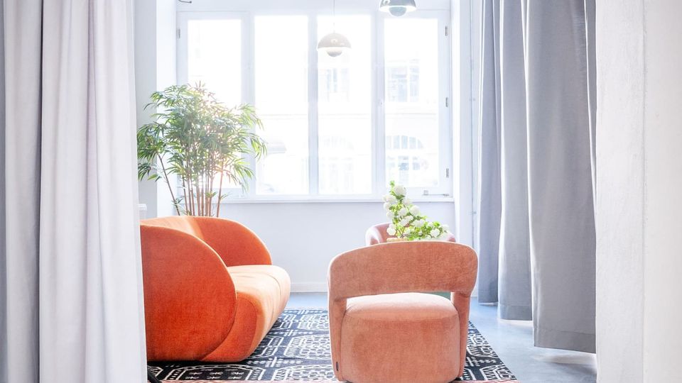 Heller, moderner Loungebereich mit großer bogenförmiger Fensterfront, grauen Vorhängen und glattem Boden. In der Mitte stehen ein orangefarbenes Sofa und ein rosafarbener Sessel auf einem gemusterten Teppich, ergänzt durch Hängelampen und Zimmerpflanzen.