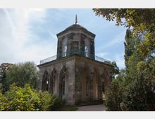 Pavillon im neogotischen Stil mit spitzbogenförmigen Arkaden, einem quadratischen Grundriss und einer Kuppel mit vergoldeter Spitze, umgeben von Bäumen und dichtem Grün. Ein Teil des Gebäudes ist von einer schmiedeeisernen Brüstung eingefasst.