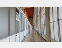 Langer, heller Flur mit großen Glasfenstern auf der rechten Seite und weißen, teilweise verglasten Wänden mit Türen auf der linken Seite. Der Boden besteht aus hellen Fliesen, und die Decke ist in einem Rotton gestrichen.